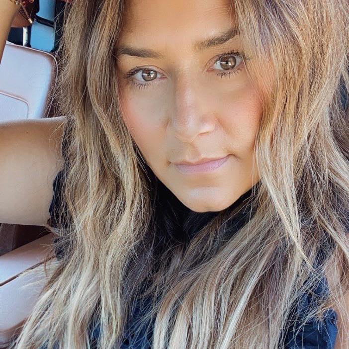 A person with long, wavy light brown hair and a neutral expression sits in a car, looking directly at the camera.