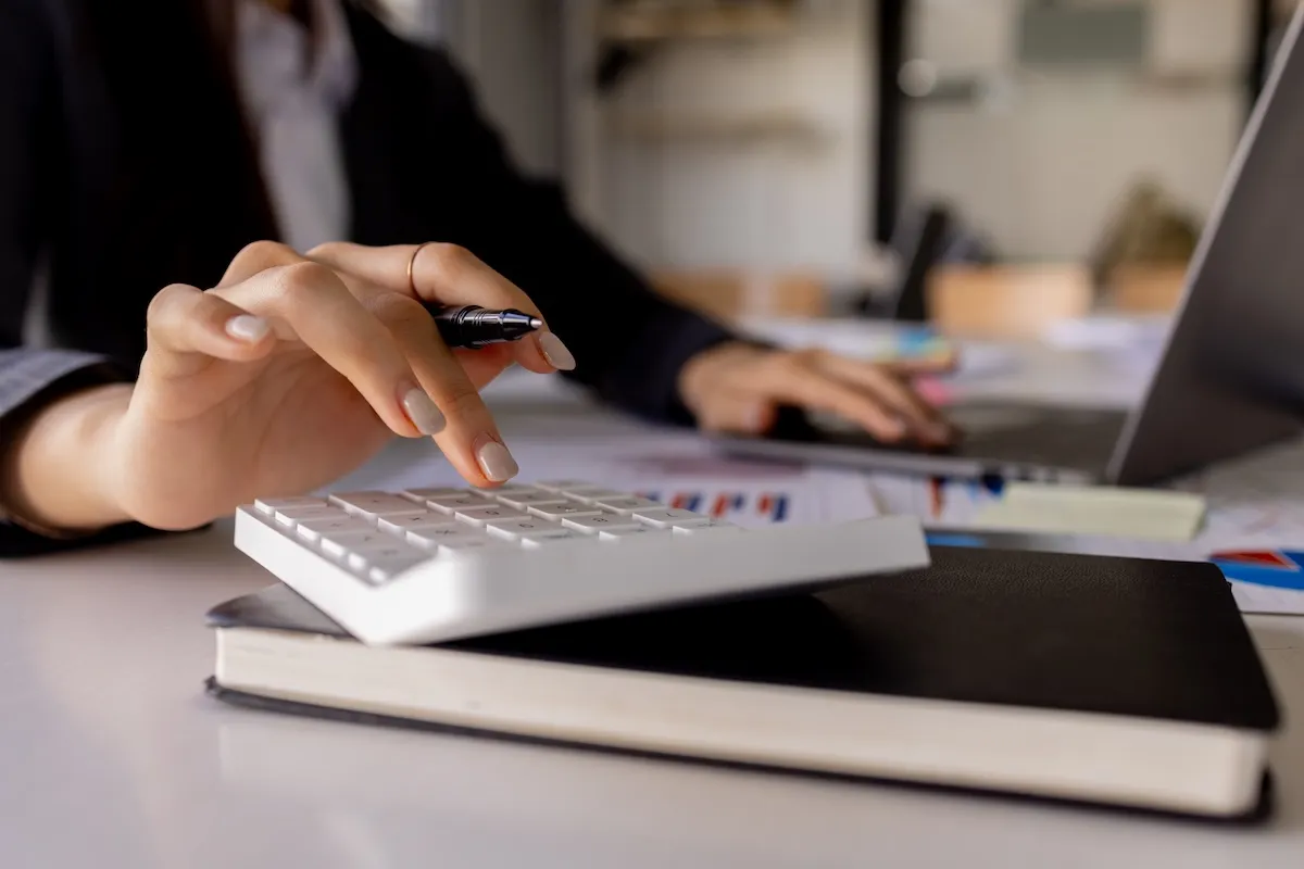 A person presses buttons on a calculator with a pen in hand, with documents, a notebook, and a laptop on the desk.