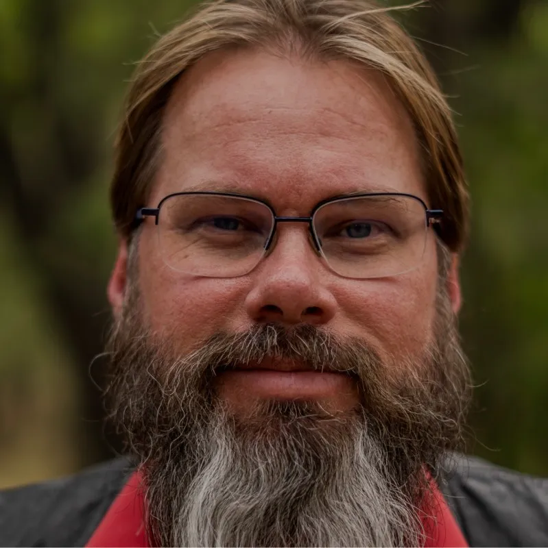 A man with glasses, a long brown and gray beard, and shoulder-length hair is facing the camera outdoors, with a blurred background.