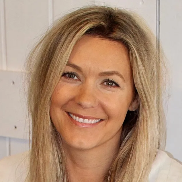 A woman with long blonde hair smiles at the camera against a light-colored wooden wall background.