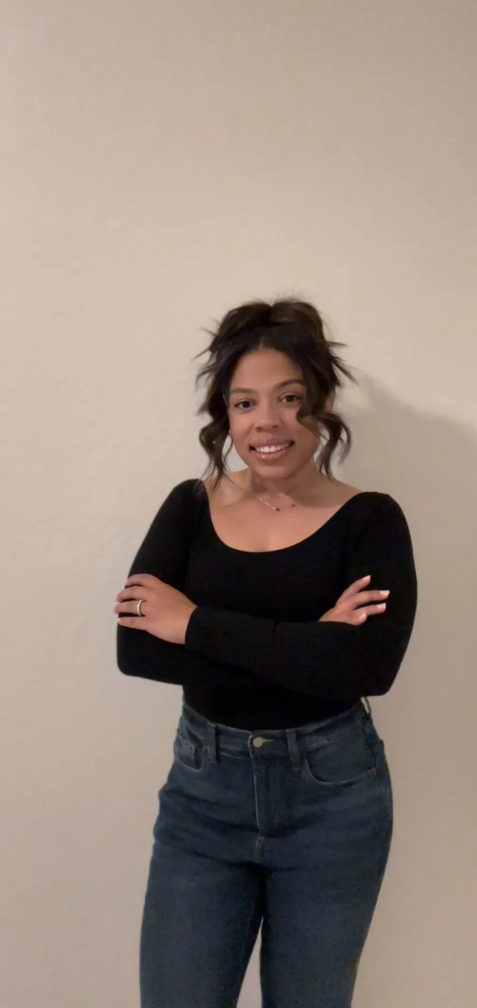 Woman with wavy hair wearing a black long-sleeve top and jeans stands against a plain beige wall with her arms crossed, smiling at the camera.