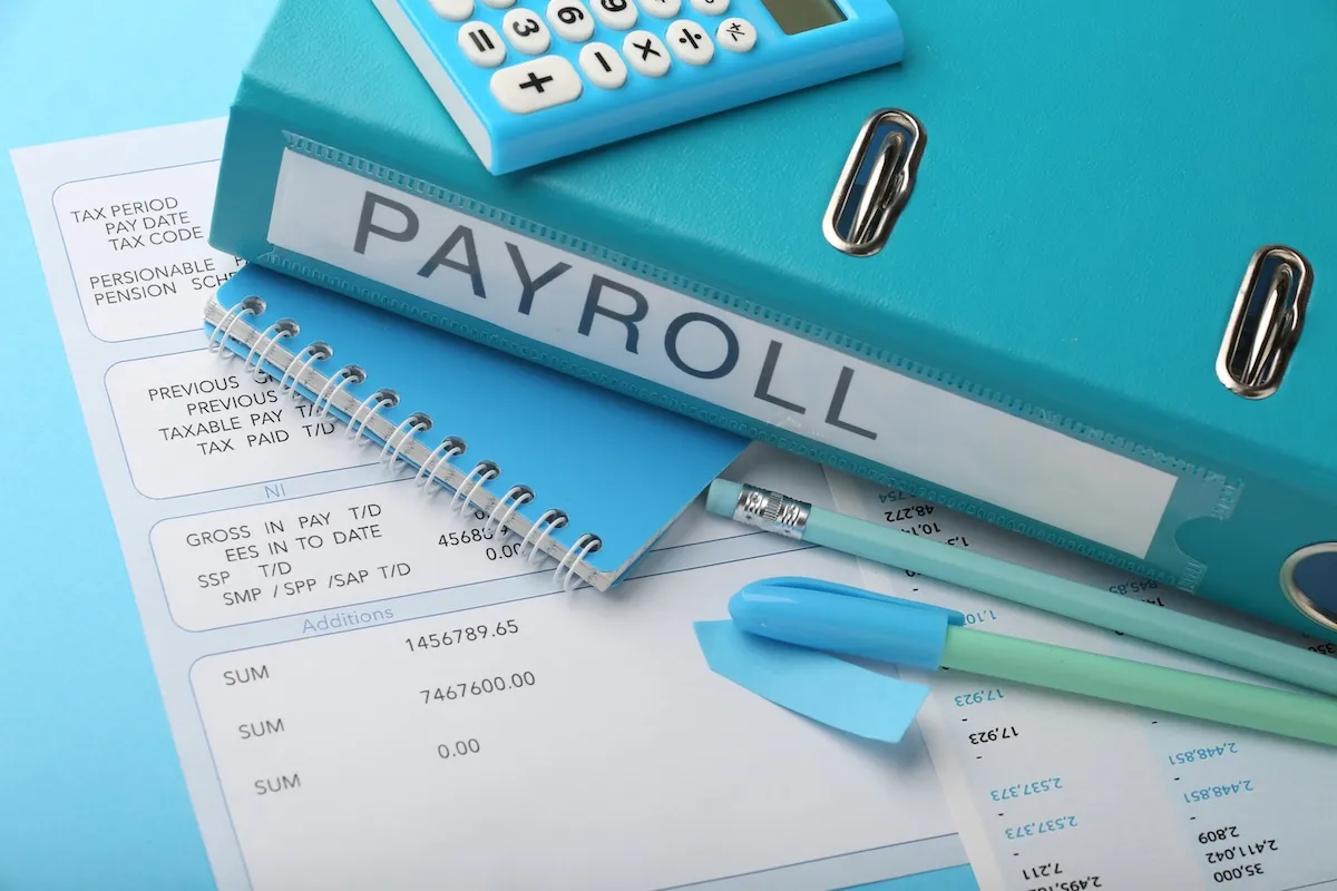 A blue payroll binder, calculator, pens, and a notepad are arranged on top of printed payroll statements.