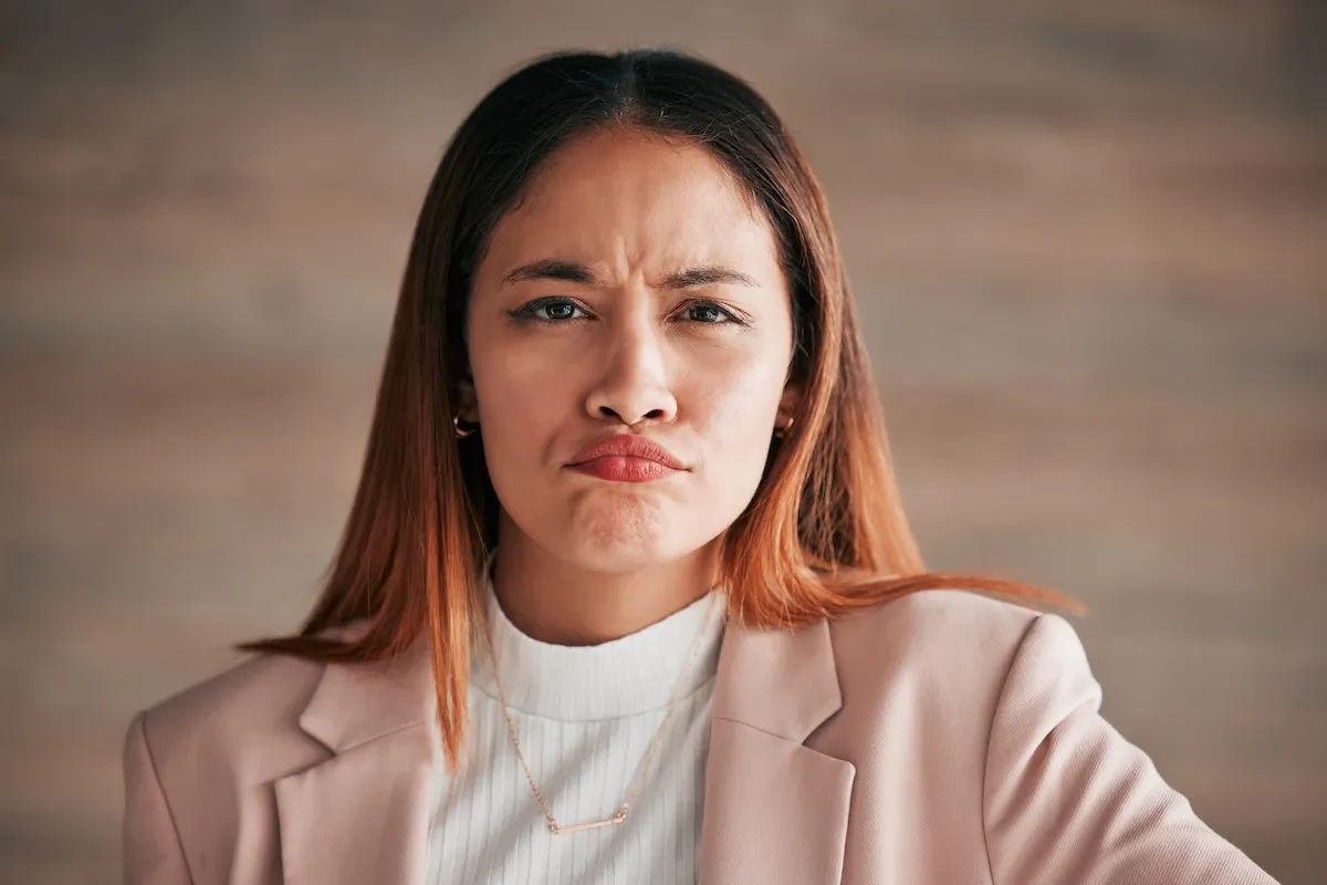 Woman with straight brown hair wearing a light pink blazer and white top, looking directly at the camera with a serious, slightly frowning expression.
