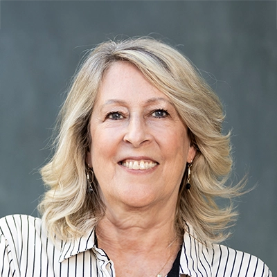 A woman with shoulder-length blonde hair, smiling, wearing a striped shirt, in front of a plain gray background.