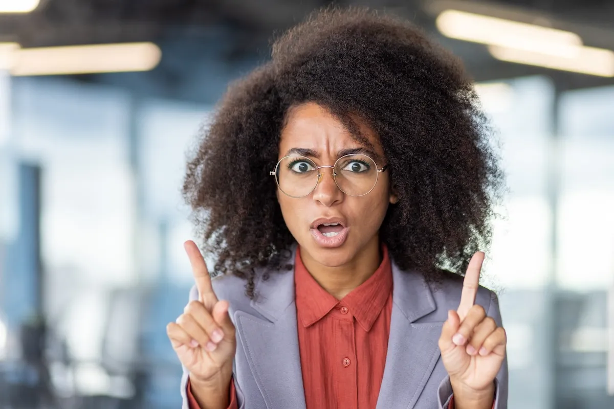 A woman with curly hair and glasses wearing a blazer and red shirt holds up both index fingers and looks surprised or concerned.