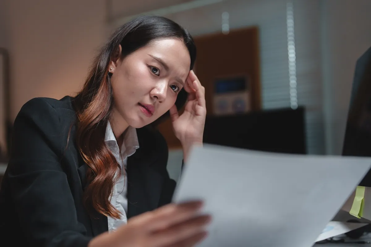 A woman in business attire sits at a desk, looking intently at a document she is holding, with a focused expression.