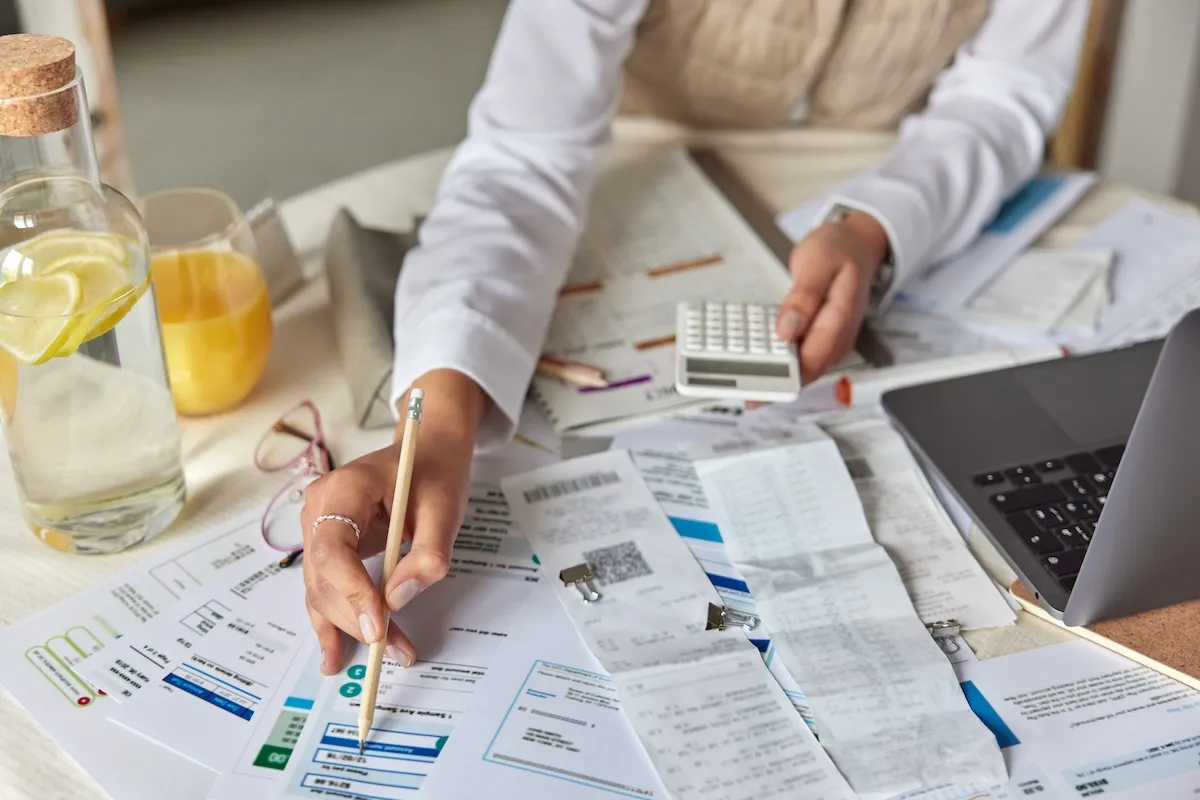 Person holding a pencil and calculator reviews bills, receipts, and financial documents on a table with a laptop, orange juice, and a jug of water with lemon.